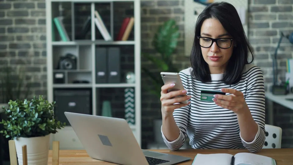 A woman at a desk with a credit card, a phone and a laptop
